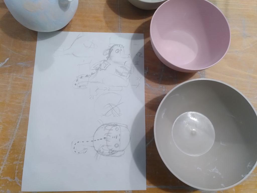 Picture of a sheet of paper with two plastic bowls of different sizes next to it.
On the sheet of paper, we can see a sketch in form of a "floor plan" and a lateral view, along with proportional dimensions.
The plan and lateral view are of a chubby dragon with tiny arms, sitting on its legs and tail and a very round head, with a huge open mouth.
The tail turns at the end as if hugging something.