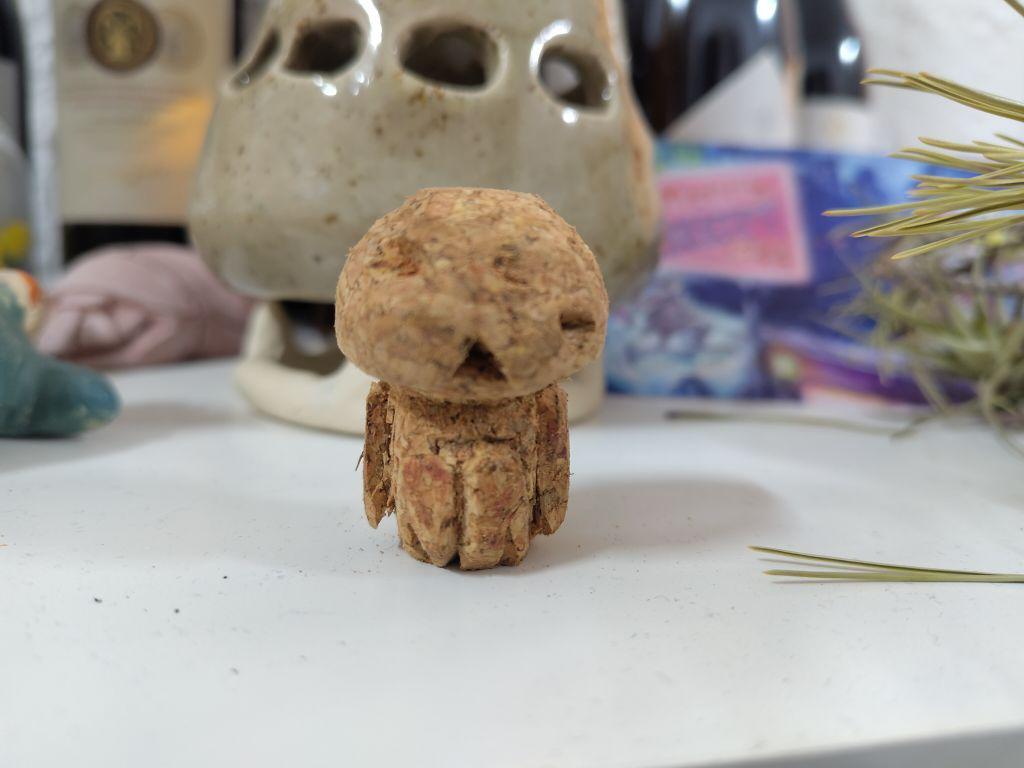 A kodama (Japanese folklore spirit) made out of a cork. It has a somewhat triangular face, with unaligned eyes and a triangular sad mouth.
