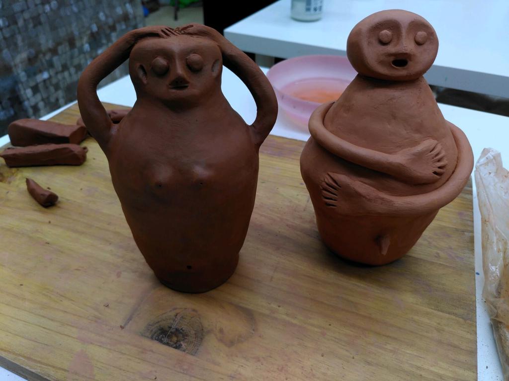 Zwei handmodellierte Figuren aus Ton. Die linke hat Bruste und Hände auf dem Kopf, die rechte armt sich um und hat ein hängendes Teil auf der unteren Seite